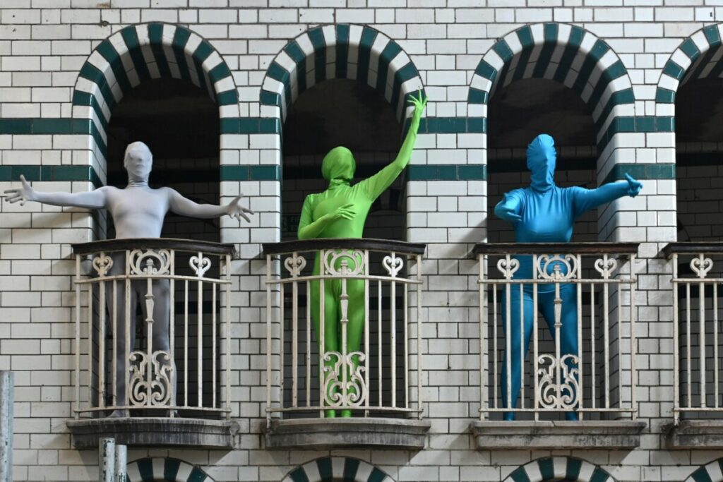Three people in morph suits stand in the Juliet balcony of Moseley Road Baths
